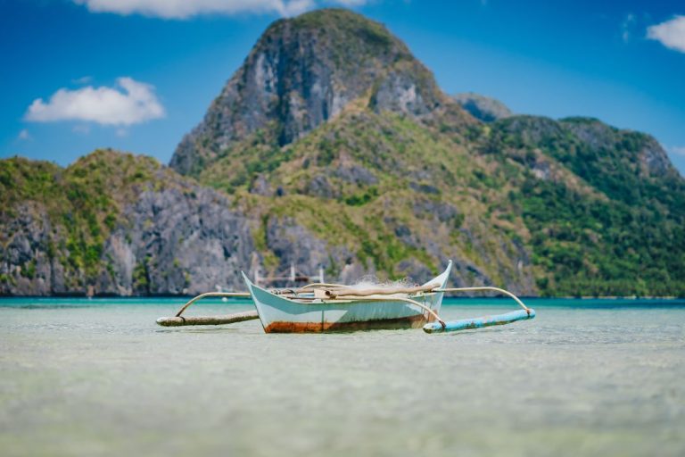 A traditional Filipino outrigger boat floats on shallow, crystal-clear water with a towering limestone cliff in the background, captured on one of the best tours in El Nido, Palawan.