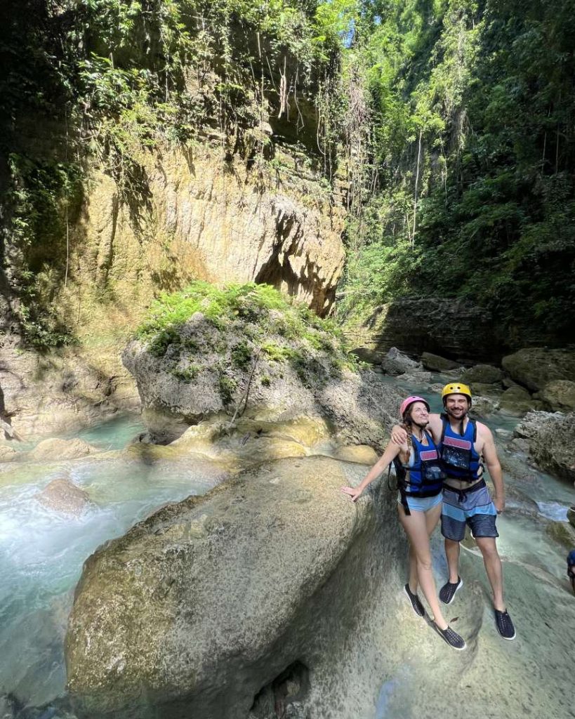 A couple wearing helmets and life vests smile while standing on a rock in a canyon stream, mid-canyoneering adventure at Kawasan Falls—an adrenaline-packed thing to do in Cebu.