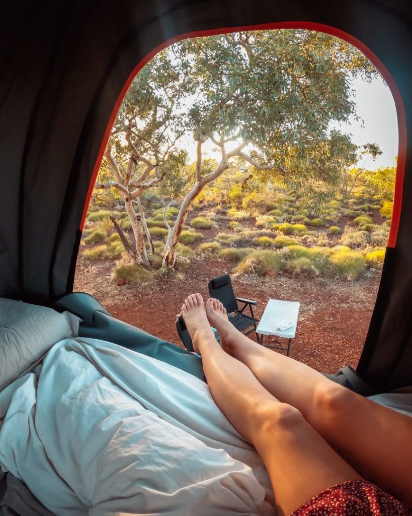 View from inside a tent of a woman’s legs stretched out towards a peaceful bush campsite in Karijini, taken during the best time to visit Karijini for camping under the stars.