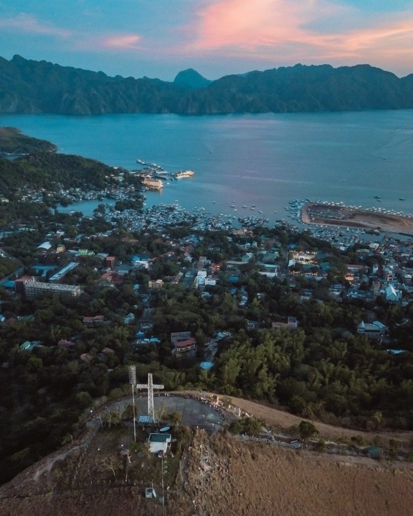 Aerial view of Coron, Philippines, at sunset, with the town nestled between lush hills and a calm bay dotted with boats. A top place to visit in the Philippines, known for its breathtaking landscapes and island-hopping adventures.