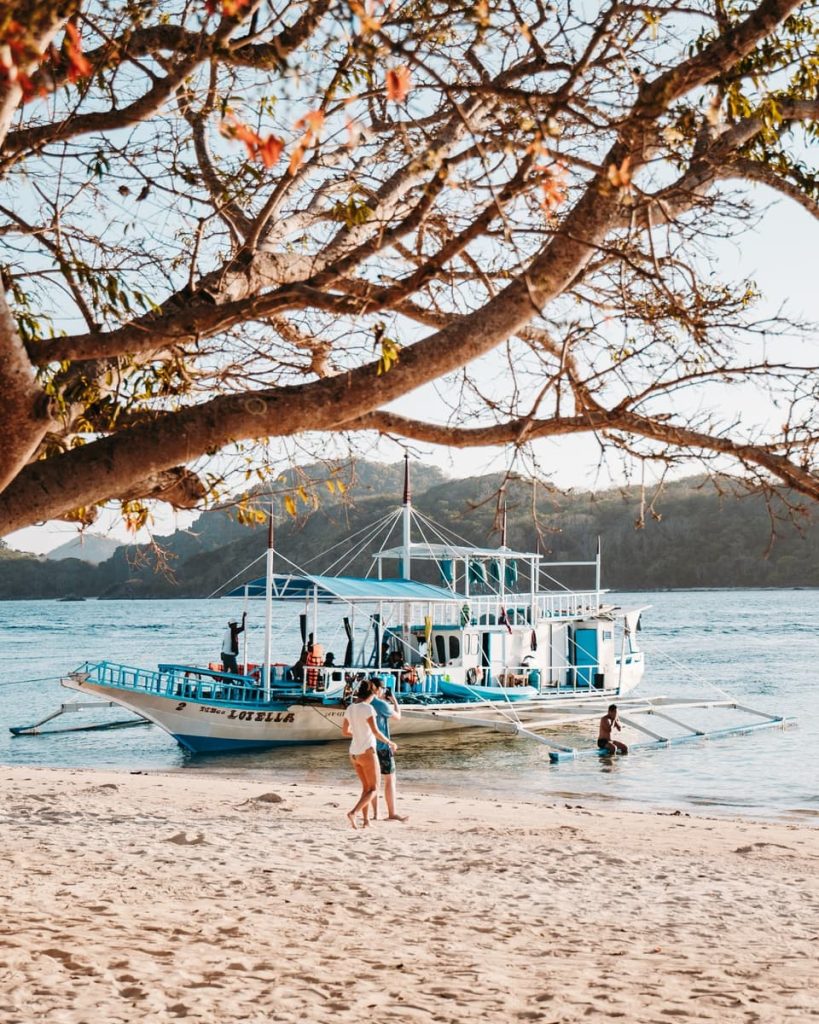 A large white boat docked at a beach under a tree’s shade, with people boarding and waving—captured during an island hopping tour from El Nido to Coron, one of the best tours in El Nido and beyond.