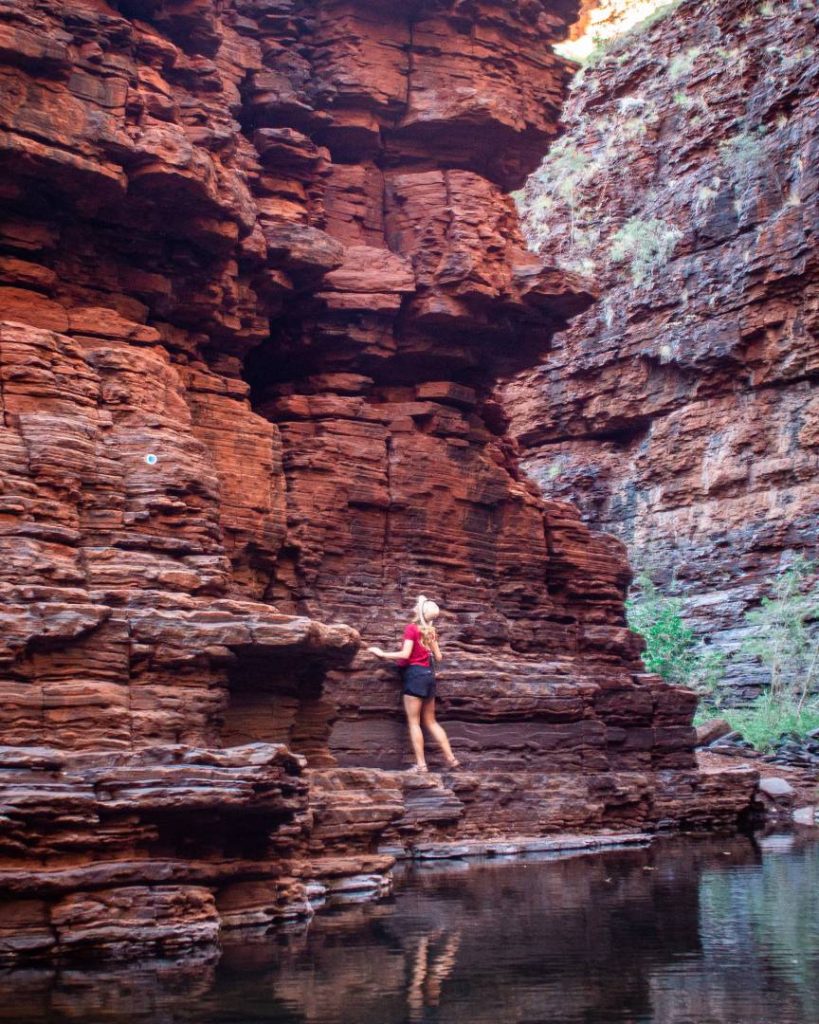 A woman climbs the rugged, layered red rock wall above a still pool in Knox Gorge, one of the best gorges in Karijini National Park. The towering cliffs rise steeply on both sides.