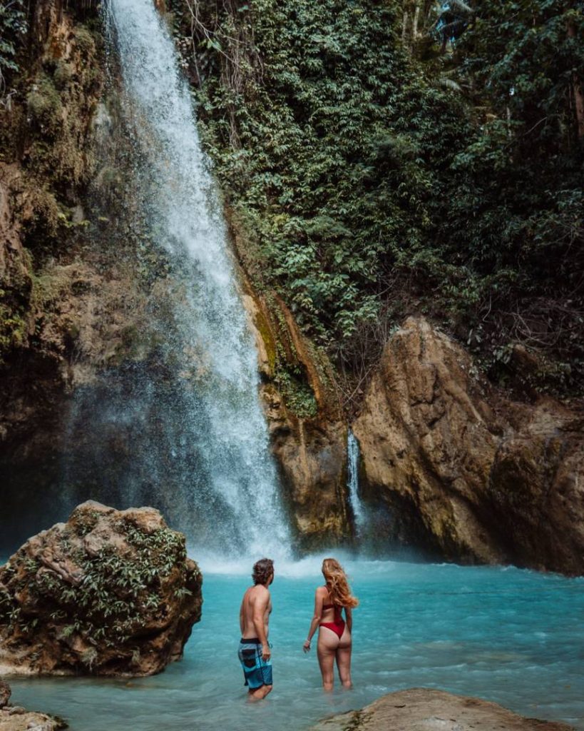 A couple standing in the turquoise waters at the base of a waterfall in Cebu, Philippines, showcasing how Philippines travel cost allows for affordable nature adventures.