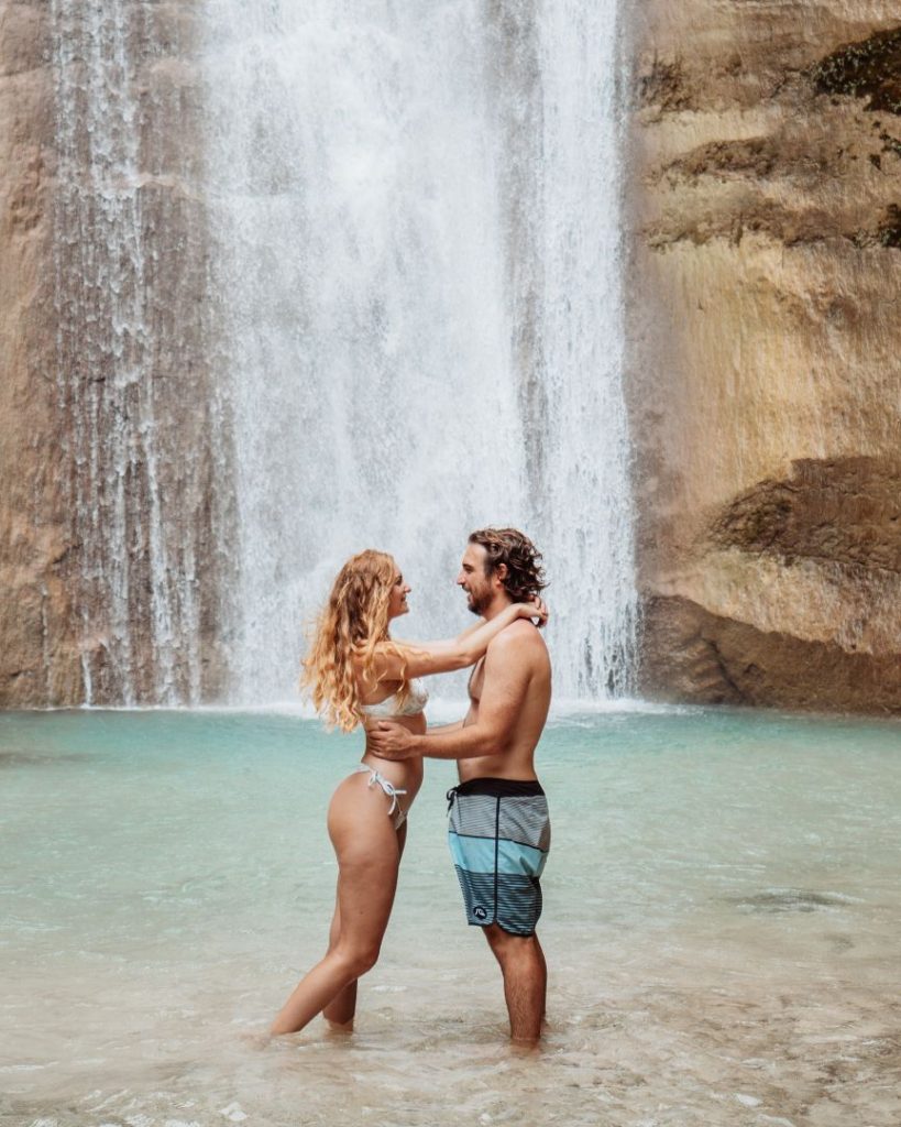 A couple stands in shallow water in front of Dao Falls, embracing under the powerful cascade—visiting this romantic spot is one of the best things to do in Cebu.