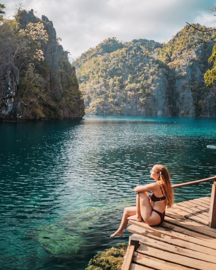 A woman sits on a wooden dock, gazing at the serene, emerald waters of Kayangan Lake in Coron, surrounded by towering limestone cliffs. One of the most beautiful spots to check out in the Philippines, offering crystal-clear waters and stunning scenery.