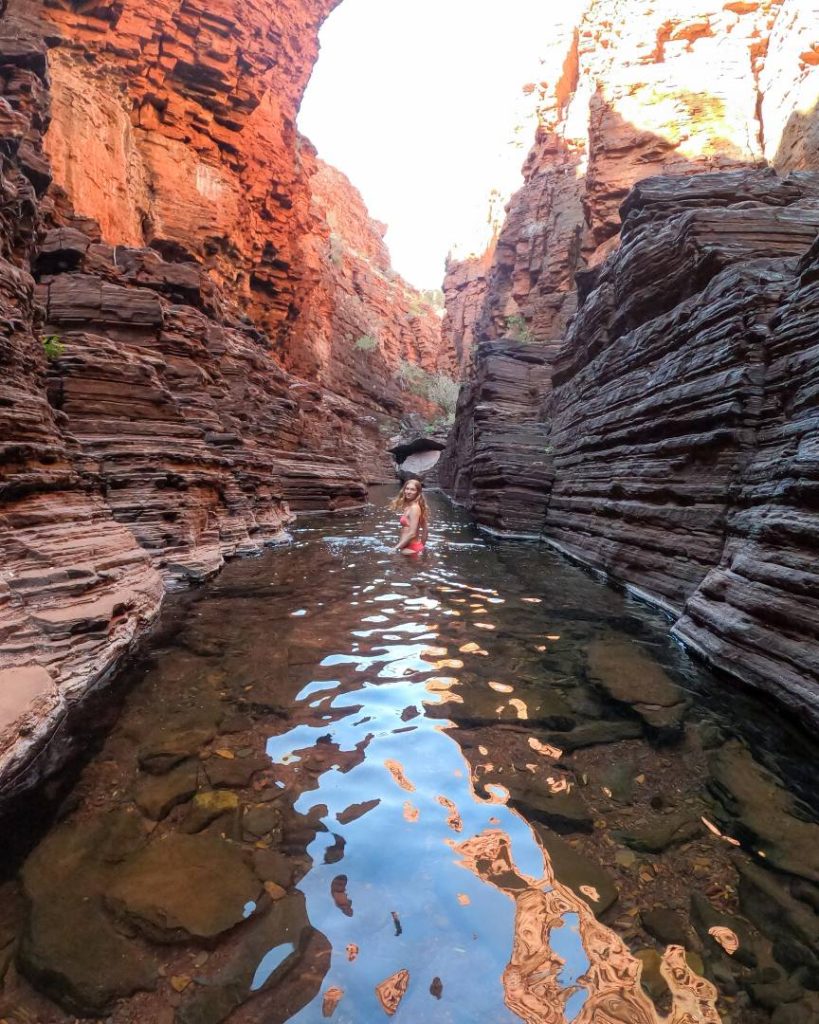 A woman wades through clear water surrounded by towering red rock walls in Knox Gorge, highlighting its spot among the best gorges in Karijini. Reflections of the cliffs shimmer in the water.