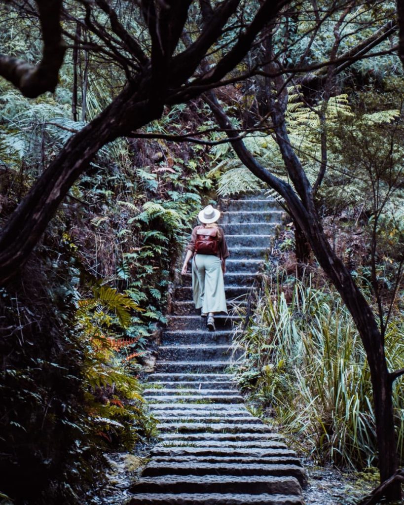 A woman walks up mossy stone steps surrounded by ferns on the way to Leura Cascades—this hike is one of the most calming things to do in the Blue Mountains.