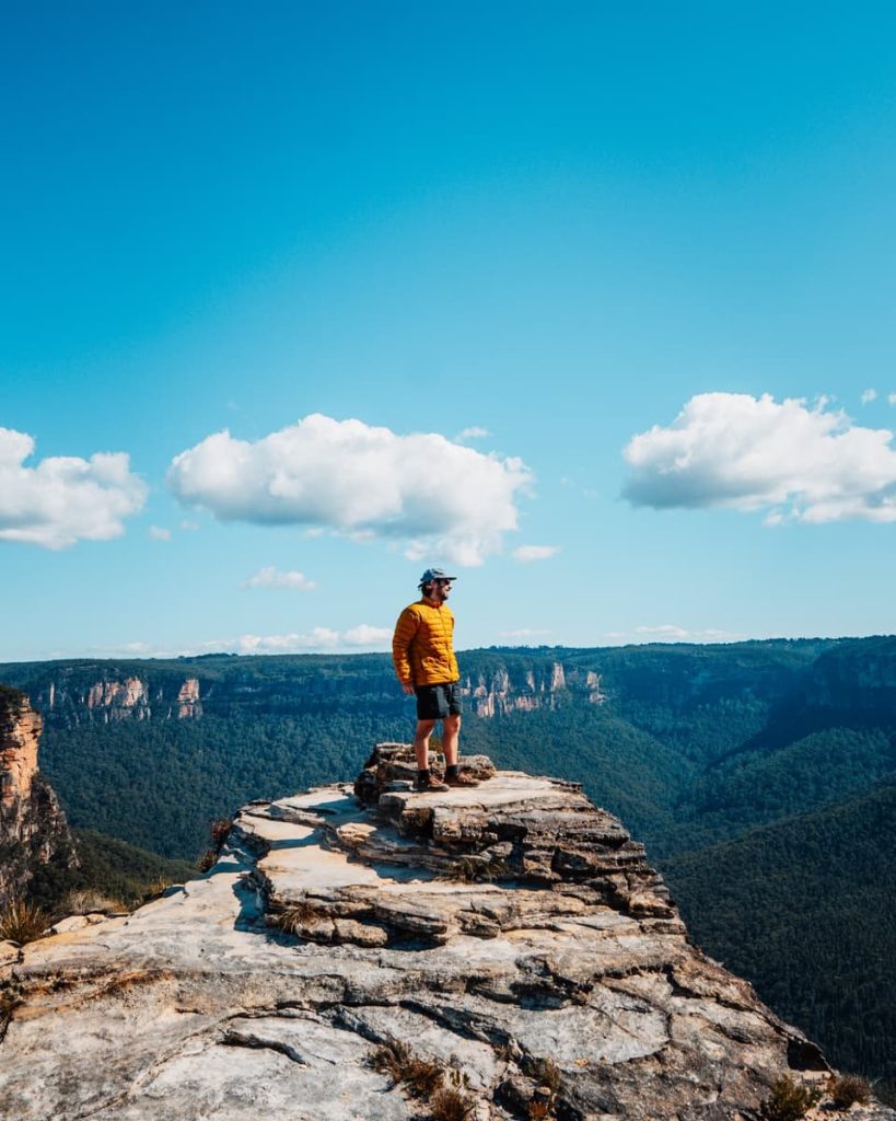 A hiker stands atop Lockleys Pylon under a wide blue sky, with sweeping valley views—an epic summit and one of the top things to do in the Blue Mountains.