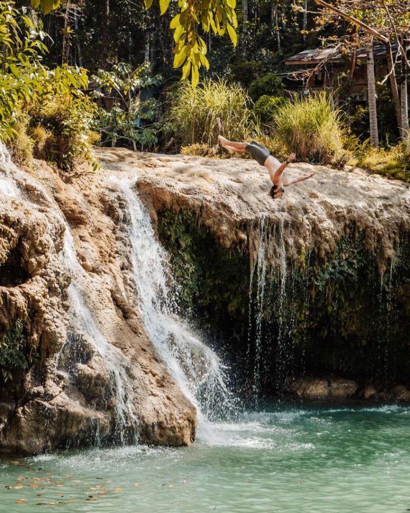 A man performs a backflip off a rocky waterfall in Siquijor, Philippines, plunging into the emerald-green pool below. A must-visit place in the Philippines, famous for its mystical charm and breathtaking waterfalls.
