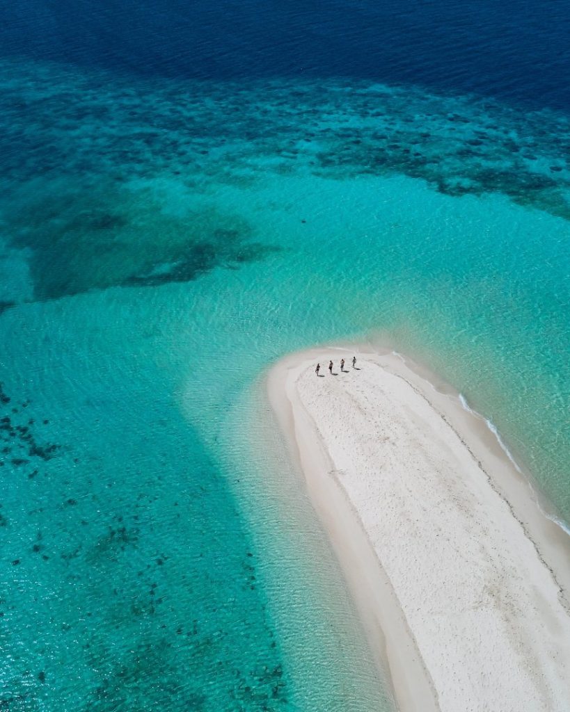 Aerial view of a remote sandbar in the Philippines, surrounded by crystal-clear turquoise waters with four people walking along the white sand. A breathtaking place to visit in the Philippines, perfect for island-hopping adventures.