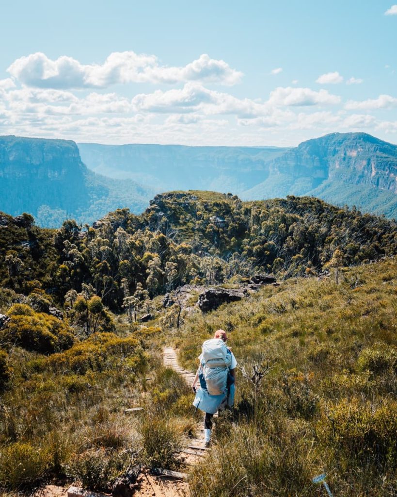 A solo hiker walks through open bushland toward Lockleys Pylon with dramatic cliffs in the distance—bushwalking here is one of the best things to do in the Blue Mountains.