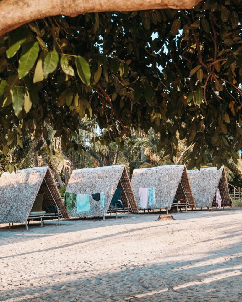 Traditional A-frame beach huts made of bamboo and nipa palm sit on a sandy shore in the Philippines, with laundry drying outside. A budget-friendly place to stay in the Philippines, offering a rustic island experience.