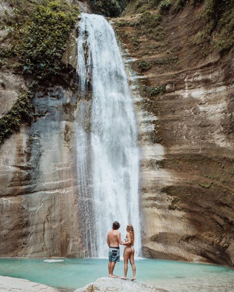 Two people stand together at the base of Dao Falls, admiring the towering waterfall that plunges into a clear blue basin—an underrated thing to do in Cebu.