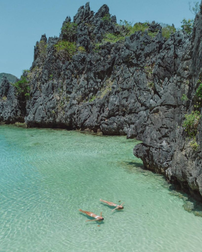 Two people float in the clear, shallow waters of Secret Lagoon, surrounded by black limestone walls—a peaceful moment in one of the best tours in El Nido.