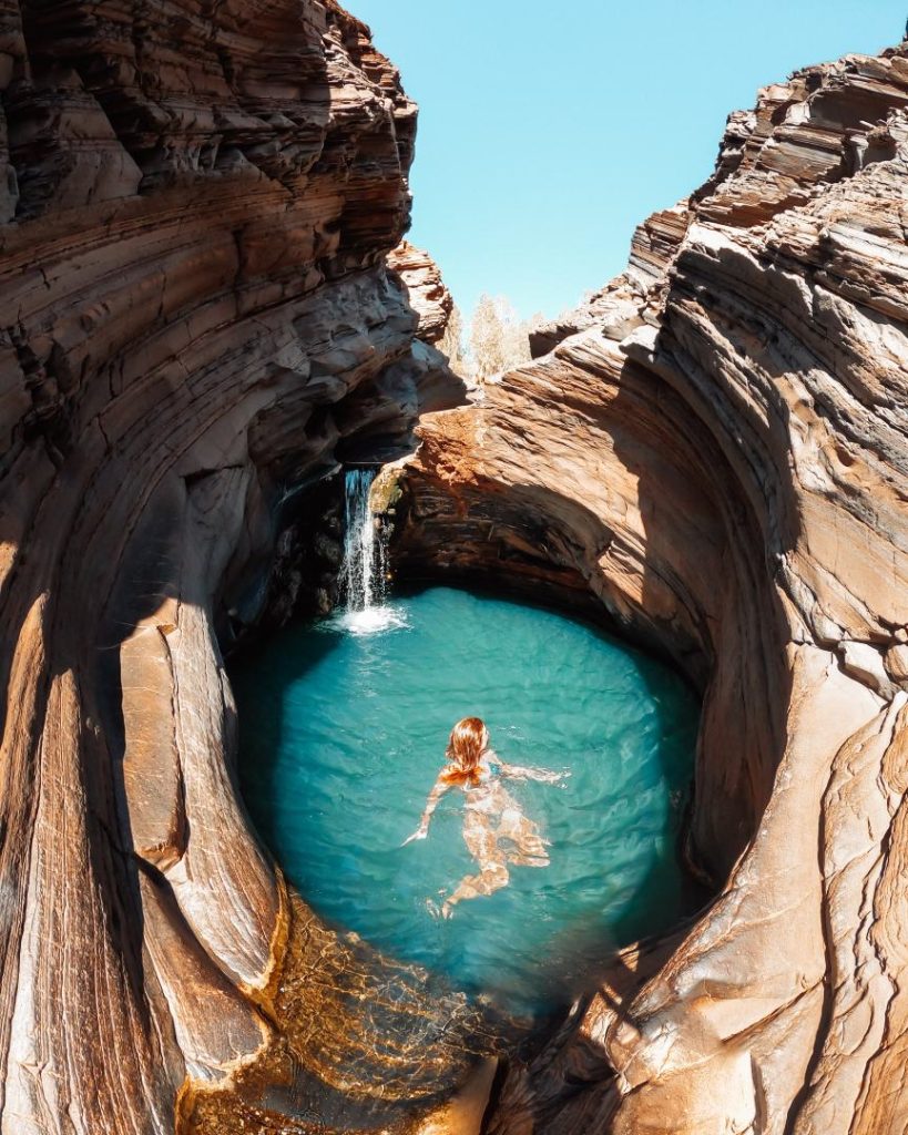 A woman swims in the turquoise waters of Spa Pool, a natural rock pool with a small waterfall and curved rock walls in Hamersley Gorge, one of the best gorges in Karijini. The sunlight highlights the layered rock textures.