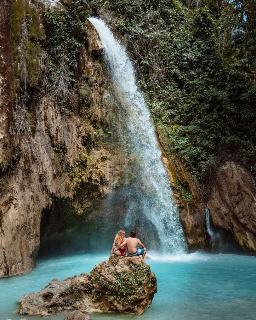 A couple sits on a rock facing Inambakan Falls, where a powerful stream of water drops into a vivid turquoise pool—one of the most scenic things to do in Cebu.