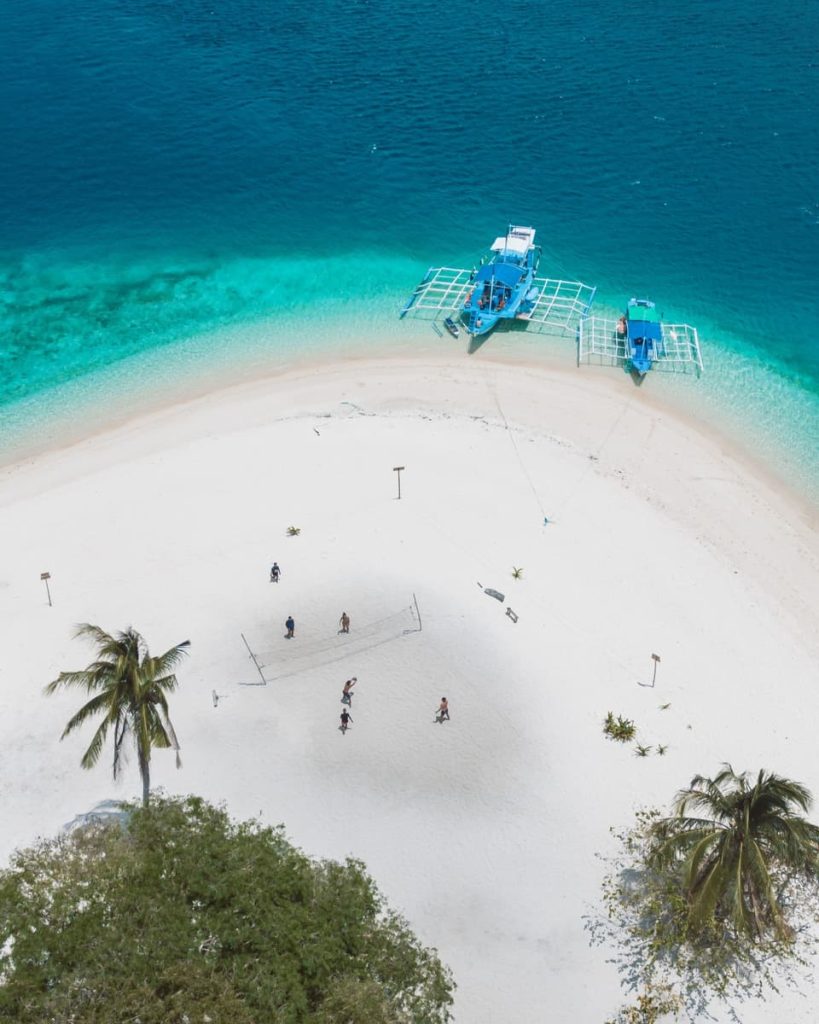 Aerial view of a white sand beach where people play volleyball, with two blue boats anchored in vibrant turquoise water—a highlight from Tour B, one of the best tours in El Nido.