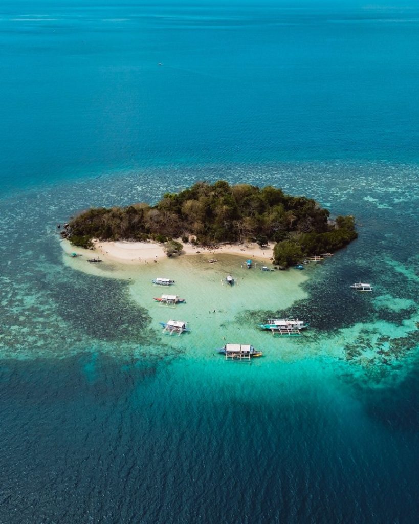 An aerial view of a small tropical island in the Philippines, surrounded by turquoise waters with several boats anchored along its sandy shoreline, illustrating how the cost of traveling the Philippines can be surprisingly low for such paradise-like destinations.
