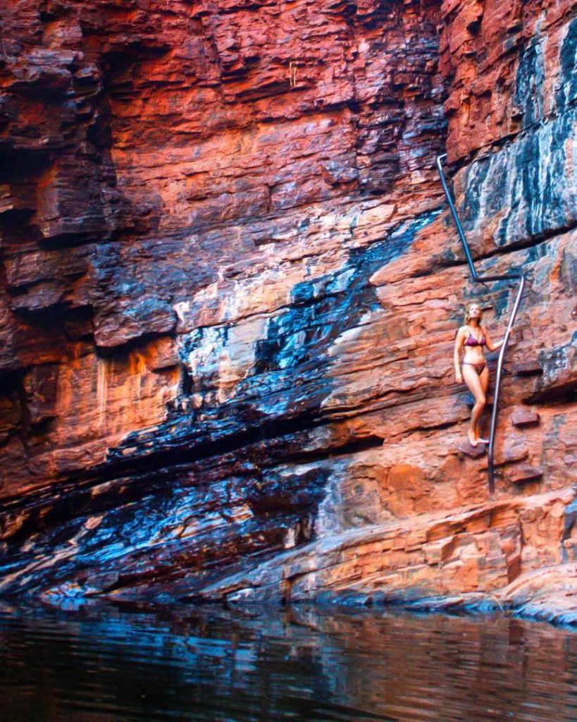 A woman descends metal handrails fixed into the rock face at Handrail Pool, surrounded by the rusty red cliffs of Weano Gorge, a must-visit among the best gorges in Karijini. Below her is dark water reflecting the cliff walls.