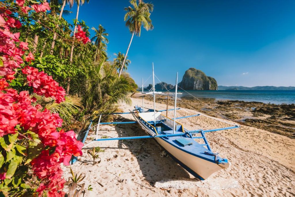 A traditional Filipino outrigger boat rests on the sandy shore of Las Cabañas Beach, El Nido, framed by lush greenery and pink bougainvillea flowers. This hidden gem in the Philippines is ideal for a relaxing beach escape.