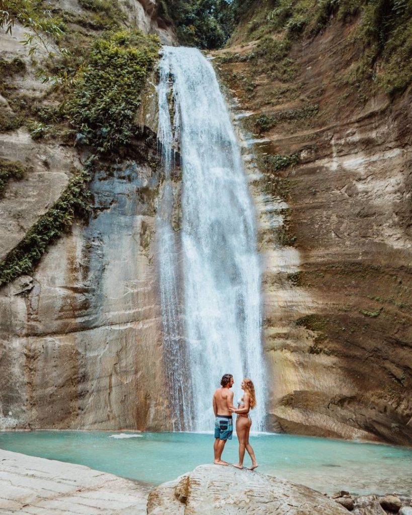 Couple standing in front of a tall waterfall with a turquoise pool, captured during the best time to visit the Philippines for outdoor adventures.