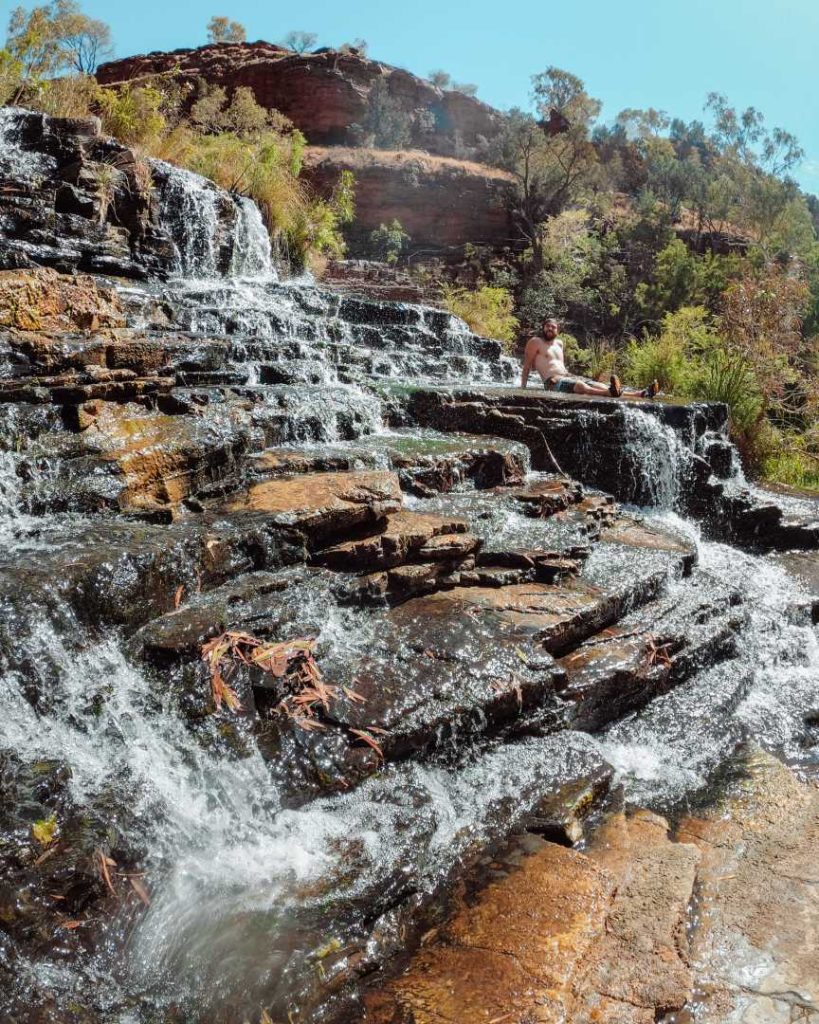 A man relaxes on the natural rock steps of Fortescue Falls, with water cascading down tiered ledges—ideal scenery during the best time to visit Karijini for swimming and exploring.