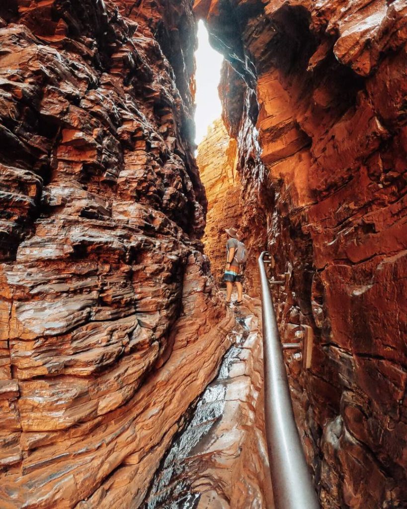 A hiker navigates a narrow passage with a handrail beside flowing water in Weano Gorge, approaching Handrail Pool in one of the best gorges in Karijini National Park, Western Australia. The orange rock walls tower above the track.