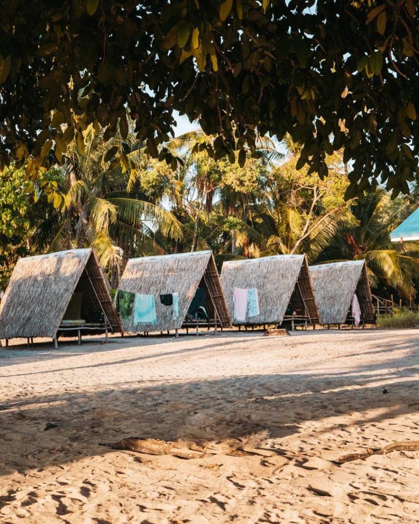 A row of thatched-roof huts on a sandy beach, with palm trees in the background, showcasing affordable accommodation options and how Philippines travel cost remains low even in paradise.