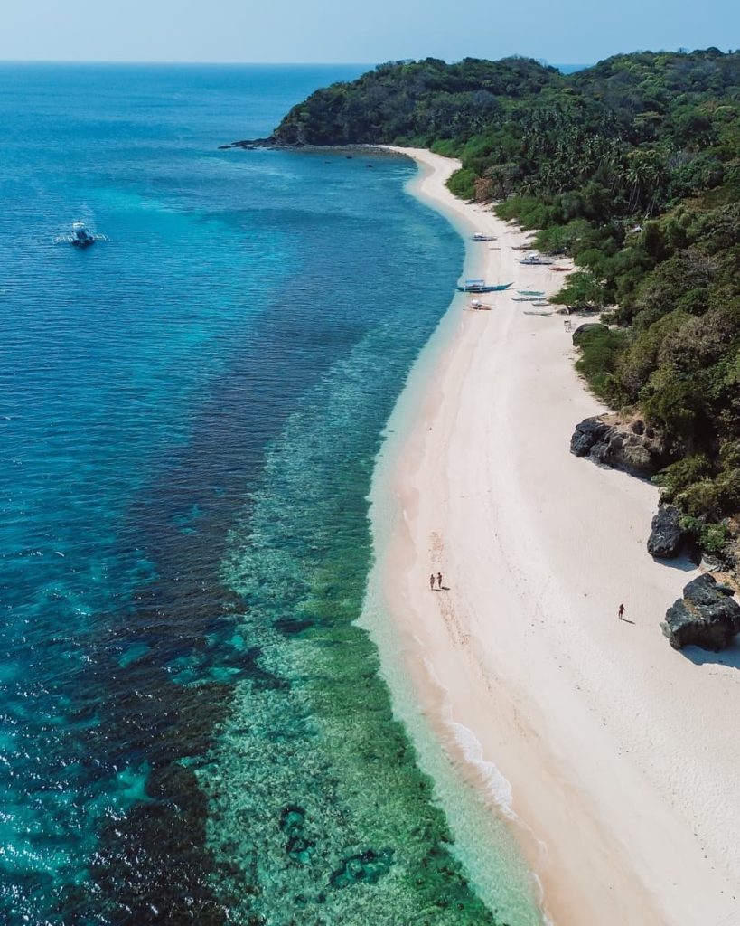 A long stretch of white sand and clear, shallow water curves around lush greenery, captured on Tour D—ideal for island hopping and one of the best tours in El Nido.