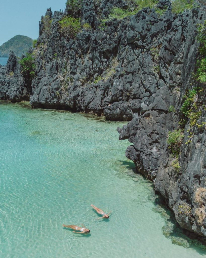 Two people float in the shallow, crystal-clear waters of Secret Lagoon, El Nido, surrounded by jagged limestone cliffs. A breathtaking place to visit in the Philippines, offering a secluded and tranquil experience.