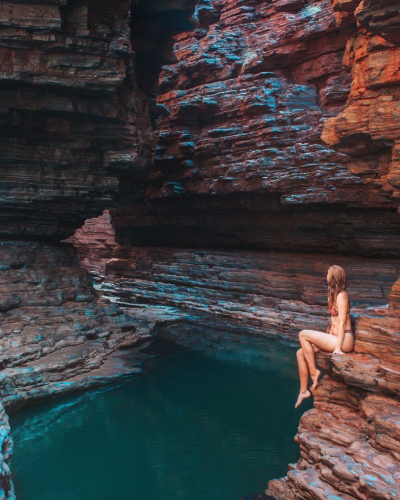 A woman in a bikini sits on a ledge above deep blue water in Kermit’s Pool, Hancock Gorge, framed by dramatic rock layers in one of the best gorges in Karijini National Park. The light highlights the smooth rock textures.