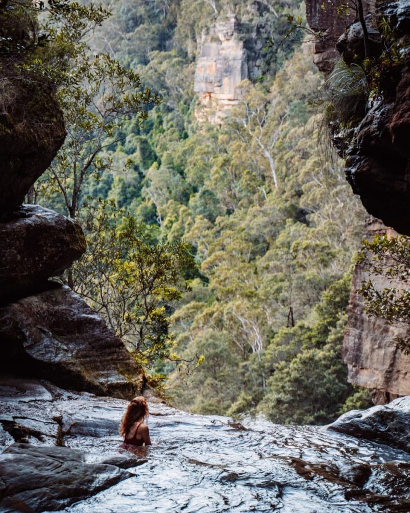 A swimmer sits in a natural rock pool at Leura Cascades with towering cliffs and forest in the distance—one of the hidden things to do in the Blue Mountains.