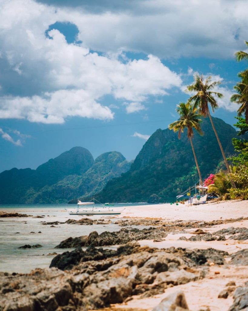 Las Cabañas Beach in El Nido features rocky shores, tall palm trees, and a distant banca boat—this scenic beach is included in some of the best tours in El Nido.