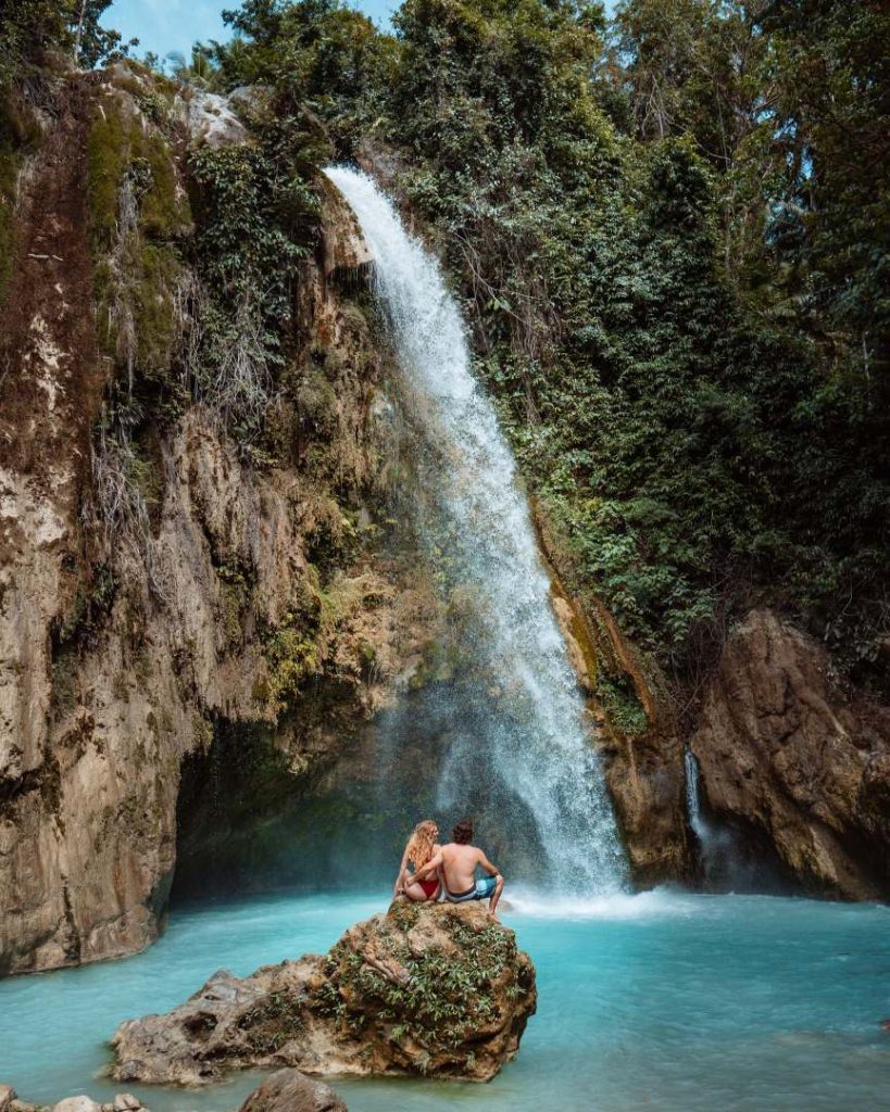 A couple sits on a rock, admiring the powerful cascade of Inambakan Falls in Cebu, surrounded by lush greenery and turquoise waters. A great place to visit in the Philippines, perfect for nature lovers and adventure seekers.