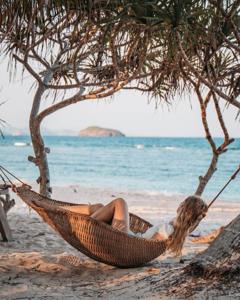 A woman relaxing in a hammock under a tree on a sandy beach, overlooking the ocean, showcasing how the cost of traveling the Philippines allows for affordable tropical escapes.