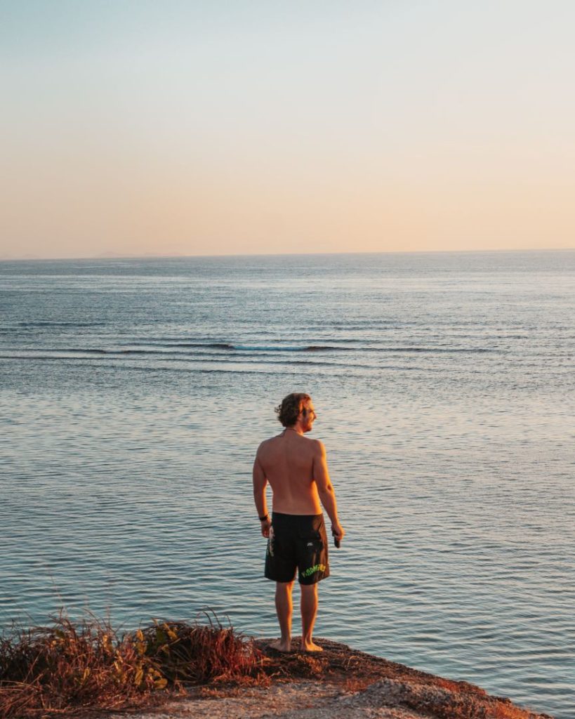 Man standing on a cliff looking out over calm ocean waters at sunset, reflecting the serenity of travelling during the best time to visit Philippines.