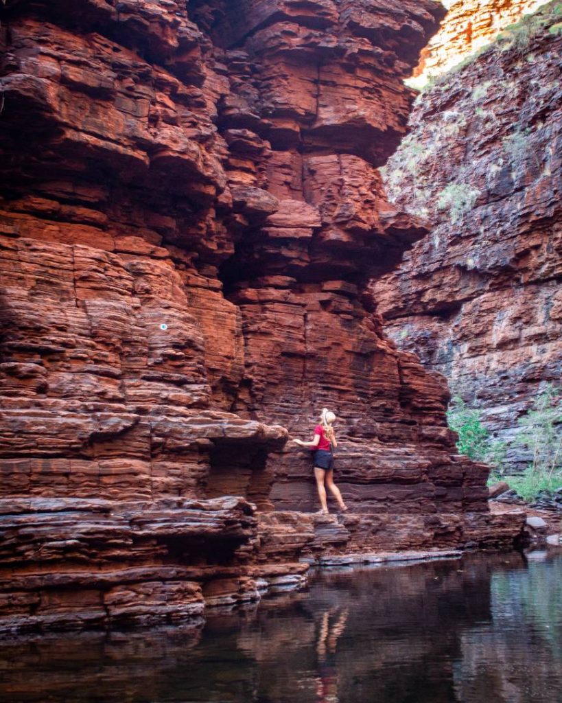 A woman in a red top carefully climbs rock ledges above water in Knox Gorge, enjoying the adventure during the best time to visit Karijini National Park.