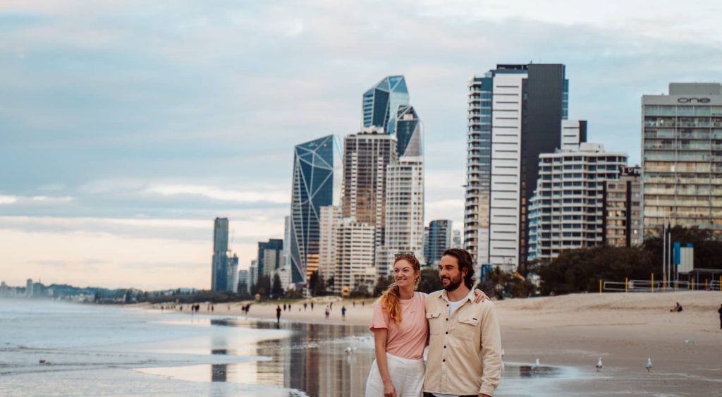 A couple stands on the wet sand of a Gold Coast beach, with the city's modern skyline towering behind them. The sky is overcast with soft, diffused light, and reflections of the buildings shimmer on the shoreline. A few beachgoers and seagulls are visible in the distance, adding to the serene coastal atmosphere.