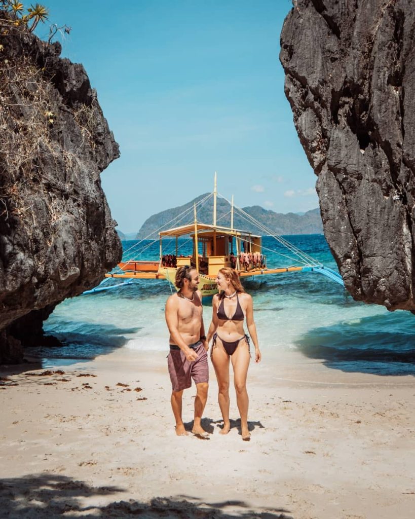 A couple walks hand in hand on a sandy beach framed by dark rock formations, with a traditional boat and island views behind them—an intimate experience from the best private boat tours in Coron.