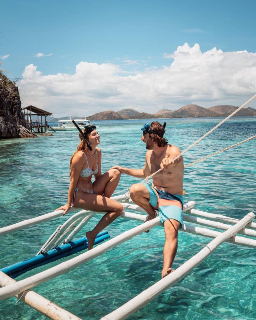 A couple in swimwear smiles at each other while sitting on the edge of a boat above clear blue waters in Coron, enjoying one of the best island hopping tours in Coron.