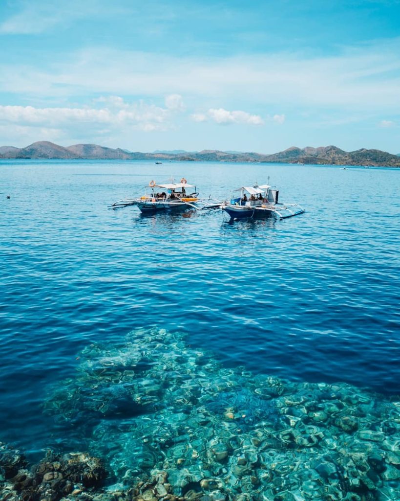 Two traditional boats float above a shallow reef in clear waters, with distant mountains on the horizon—classic scenery on the best shared boat tours in Coron, Philippines.