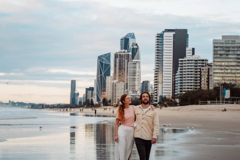 A couple walks along a beach at sunset with Gold Coast's modern skyscrapers and high-rise buildings in the background, reflected on the wet sand.