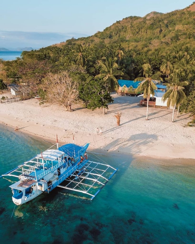 A blue boat anchored near a sandy beach lined with palm trees and small cottages, part of a Keeloma island hopping tour linking Coron and El Nido—one of the best boat tours in the Philippines.