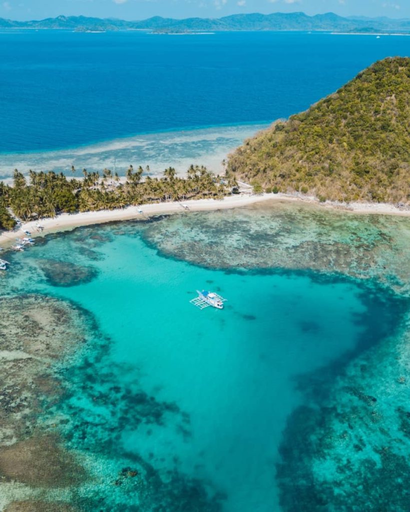 Bright turquoise reef and open sea surround a white outrigger boat near a palm-lined island on the Keeloma route, showcasing why this is one of the best island hopping tours in Coron.