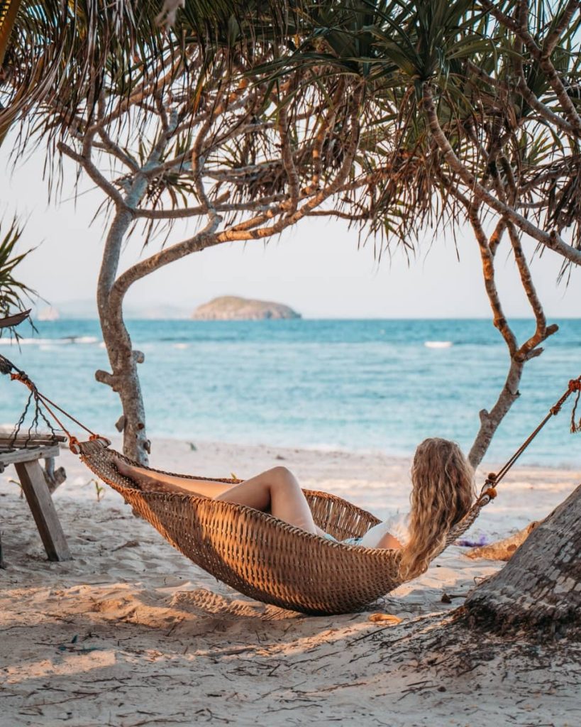 A woman lounges in a woven hammock under trees on a sandy beach with ocean views, capturing the laid-back vibe of relaxing boat tours in Coron.