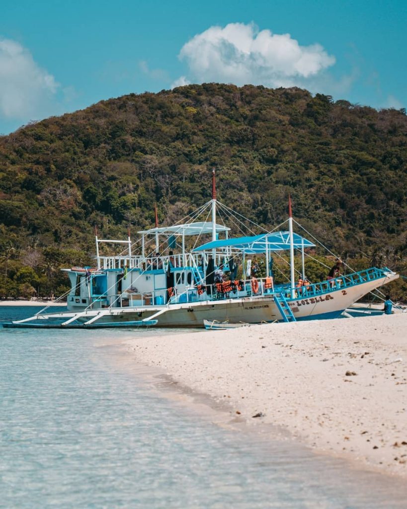 A traditional outrigger boat anchored on a quiet white-sand beach with a forested hill in the background—this was a Keeloma island hopping tour in Coron, Philippines.