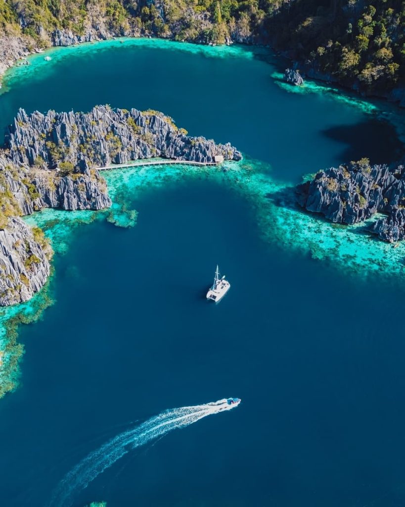 Aerial view of Twin Lagoon's deep blue water surrounded by jagged limestone cliffs, with boats gliding through—iconic scenery on the best tours in Coron.