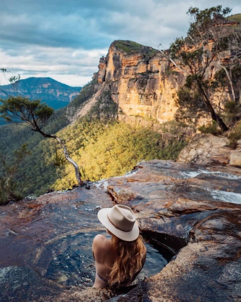 A woman in a wide-brimmed hat relaxes in the natural infinity pool at Fortress Falls, gazing out over rugged cliffs and forested valleys—an iconic spot in any Blue Mountains weekend itinerary.