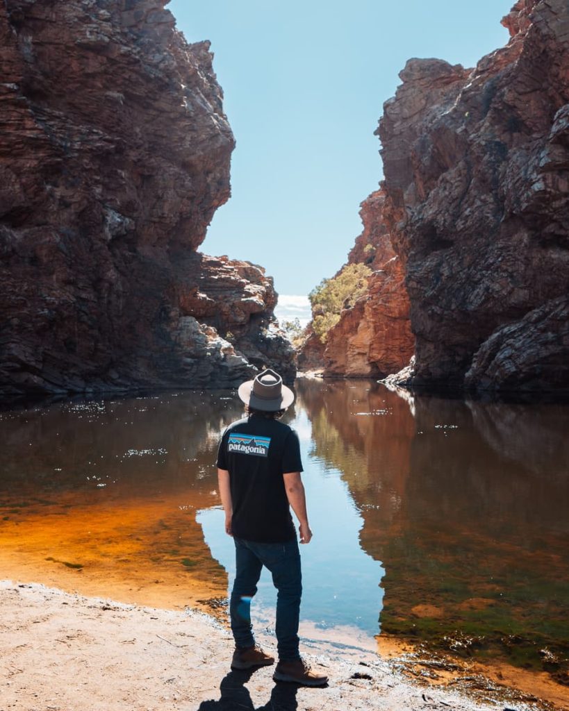 A traveller stands at the edge of Ellery Creek Big Hole in the Northern Territory, surrounded by towering red cliffs—an outback icon on road trips in Australia.