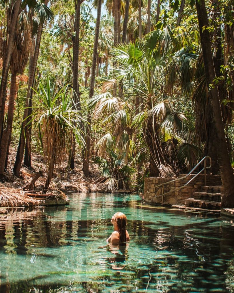 A woman relaxes in the crystal-clear waters of Mataranka Springs, surrounded by lush palms—a tranquil stop on Northern Territory road trips in Australia.